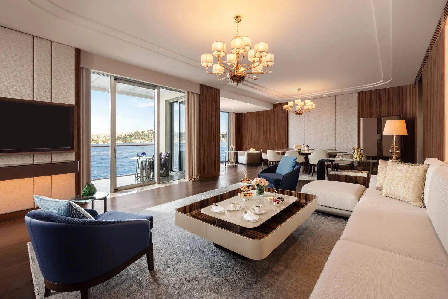 Sala de suite presidencial con sofá beige, butaca azul, mesa de centro en madera de ébano y candelabro dorado con vista al mar