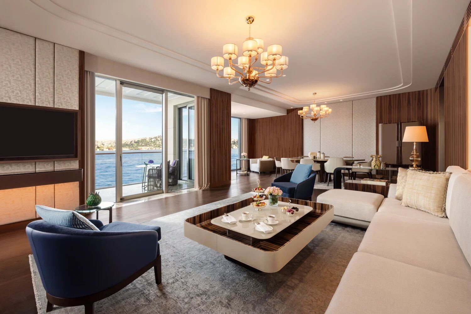 Sala de suite presidencial con sofá beige, butaca azul, mesa de centro en madera de ébano y candelabro dorado con vista al mar